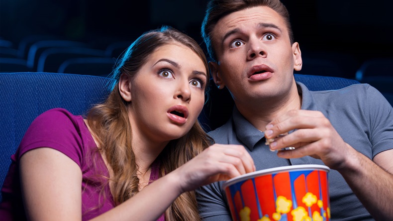 Shocked young couple eating popcorn and watching movie together while sitting at the cinema