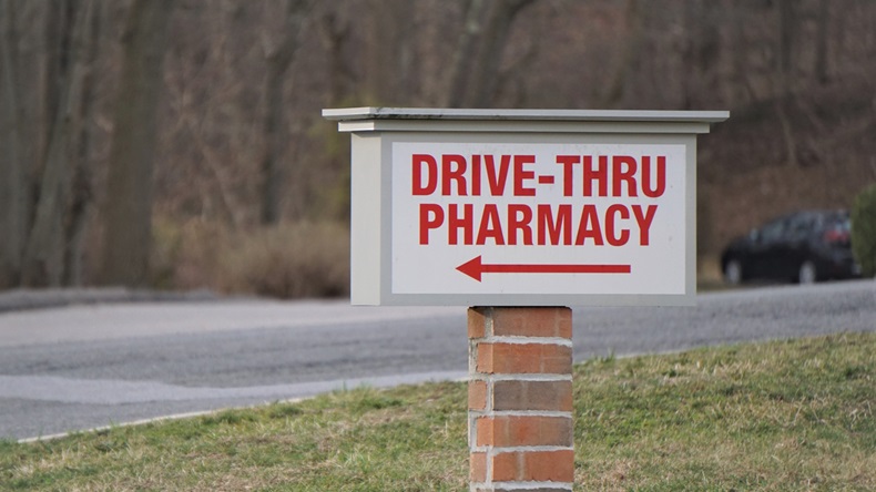 Isolated sign advertising 'Drive-Thru Pharmacy' in capitals font directs drivers with arrow. Signage on brick pedestal grass empty road winter trees in background